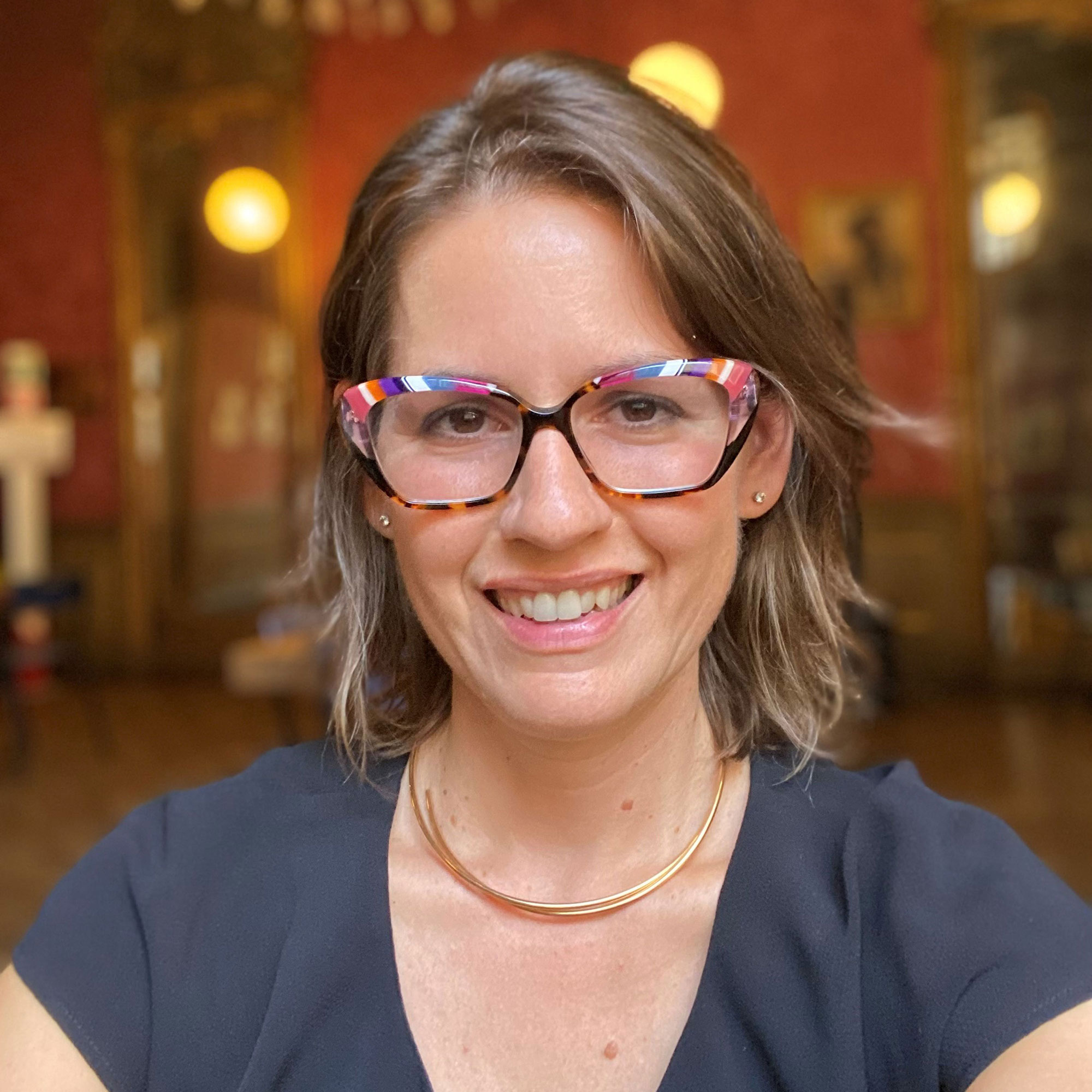 A woman wearing colorful glasses and a gold necklace smiles.