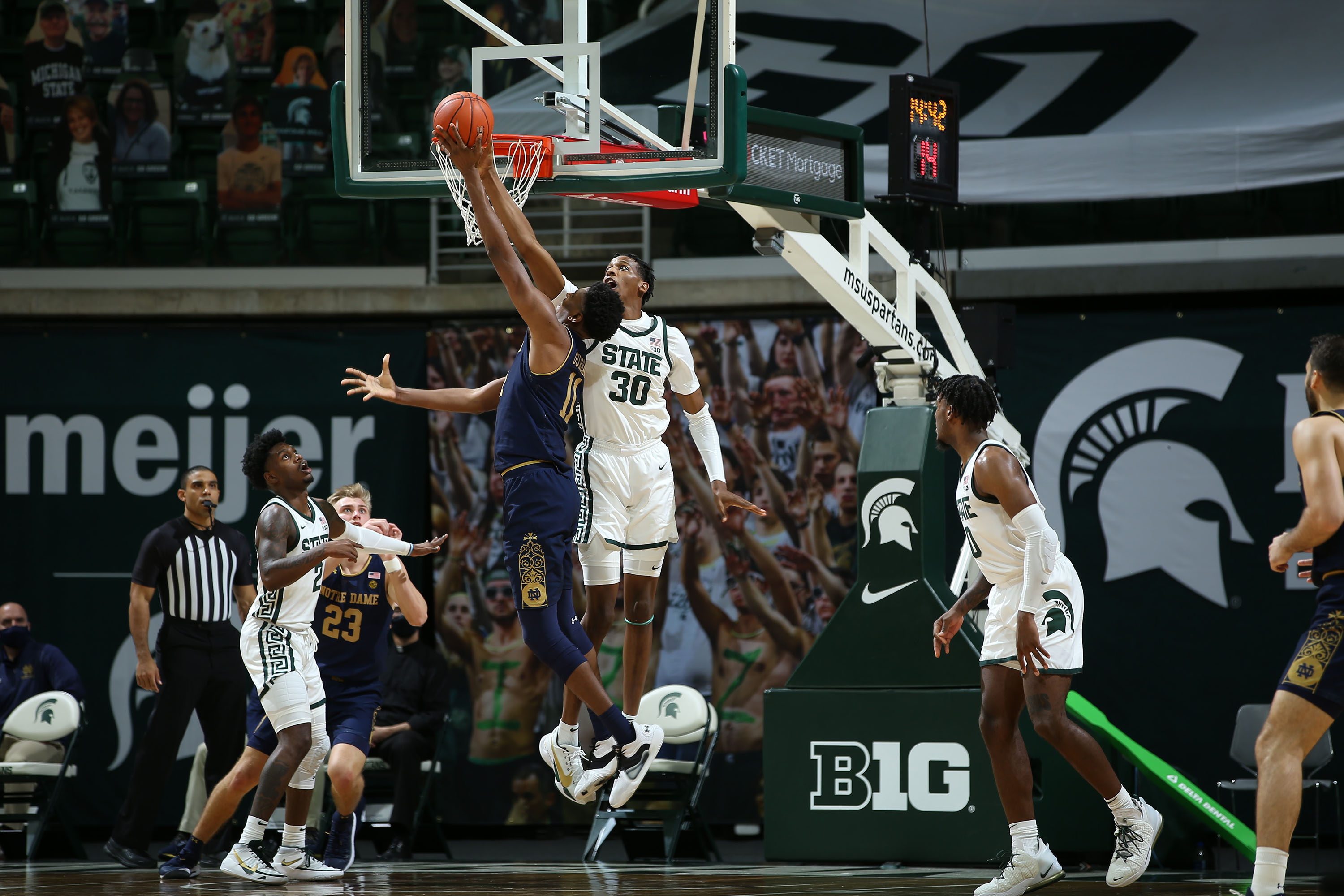 A Spartan jumps to block the shot at the rim of a Notre Dame player while two other Spartans and one Notre Dame player look on.