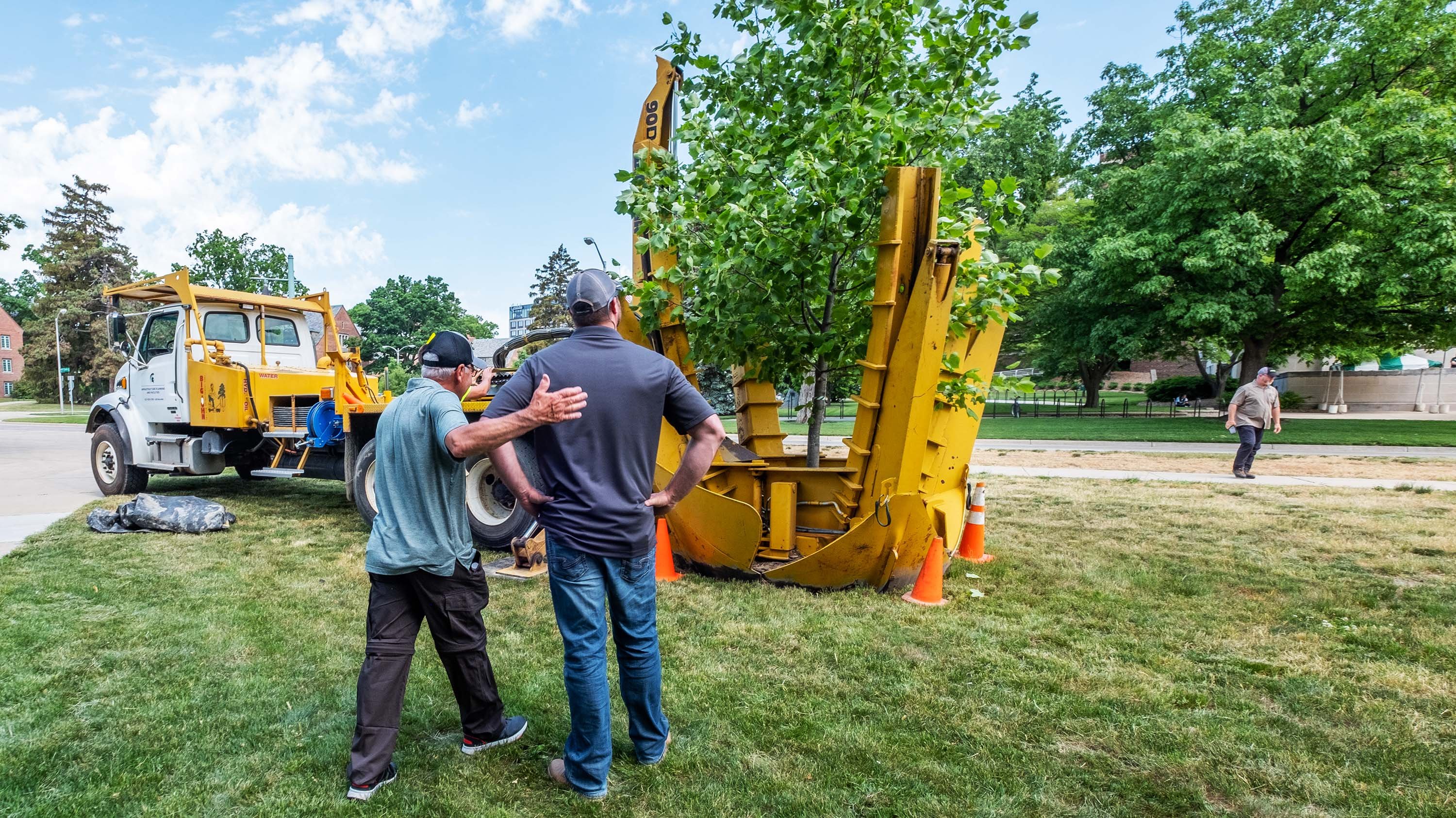 Two people, including Paul Swartz, watching as a tree is planted with a piece of machinery.