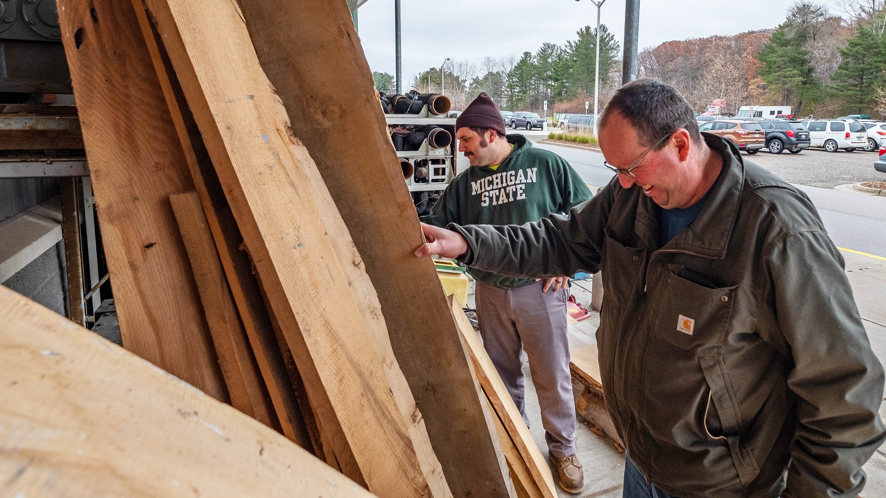 Two people look at boards of wood in front of the MSU surplus store