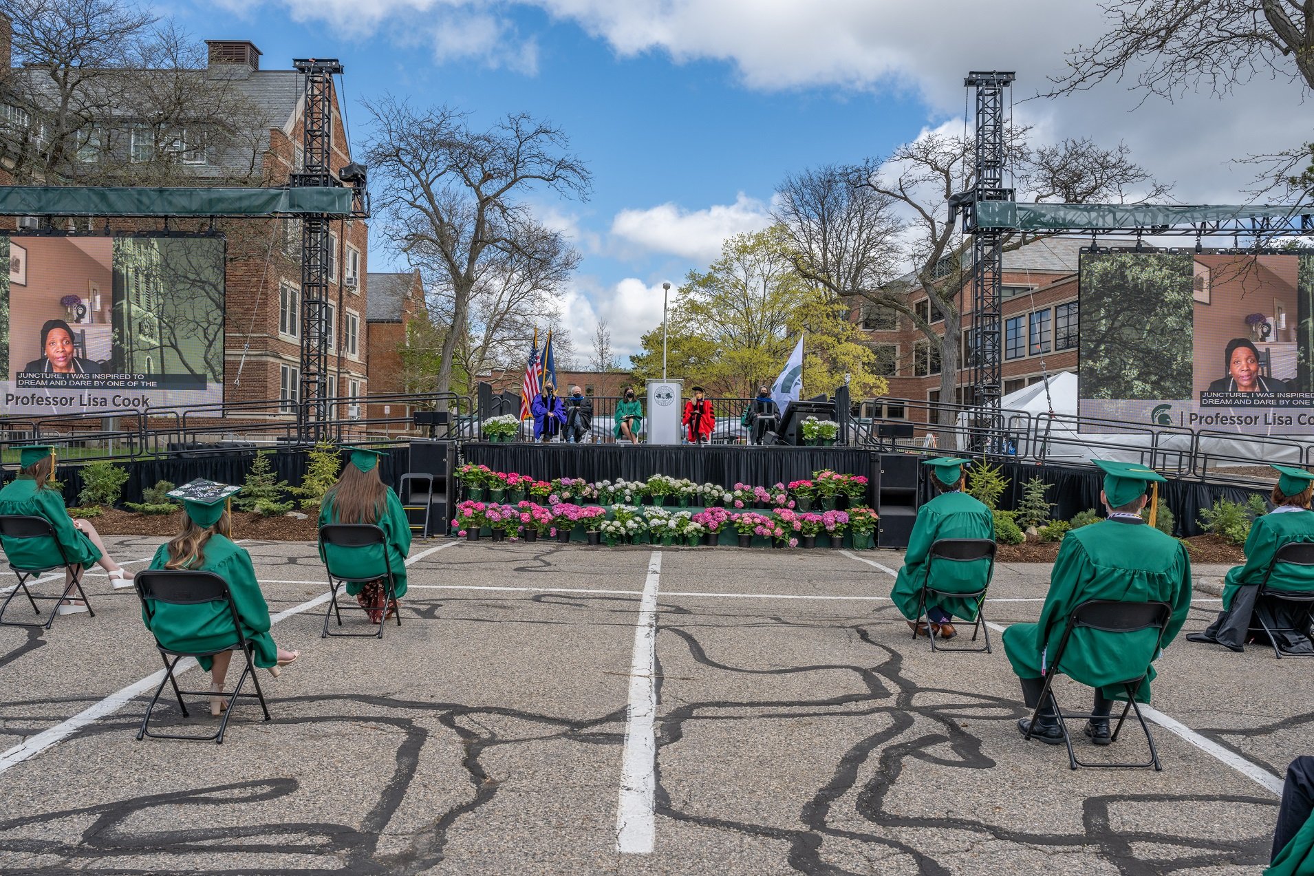 View over the shoulders of graduates in green caps and gowns at the commencement platform in the MSU Auditorium parking lot. On a platform at the front, there are seated speakers and a podium. On either side of the platform are large screens showing a video speech from Professor Lisa Cook.