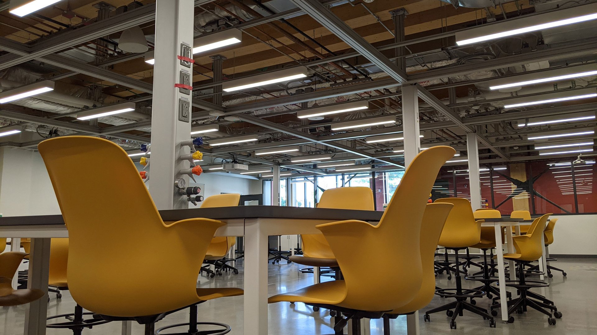 A photograph from inside the new STEM Teaching and Learning facility shows a lab space with yellow chairs, movable tables and columns mounted to the ceiling that carry air, natural gas and vacuum lines to workstations. These columns can also be moved when educators need to reconfigure their teaching space.