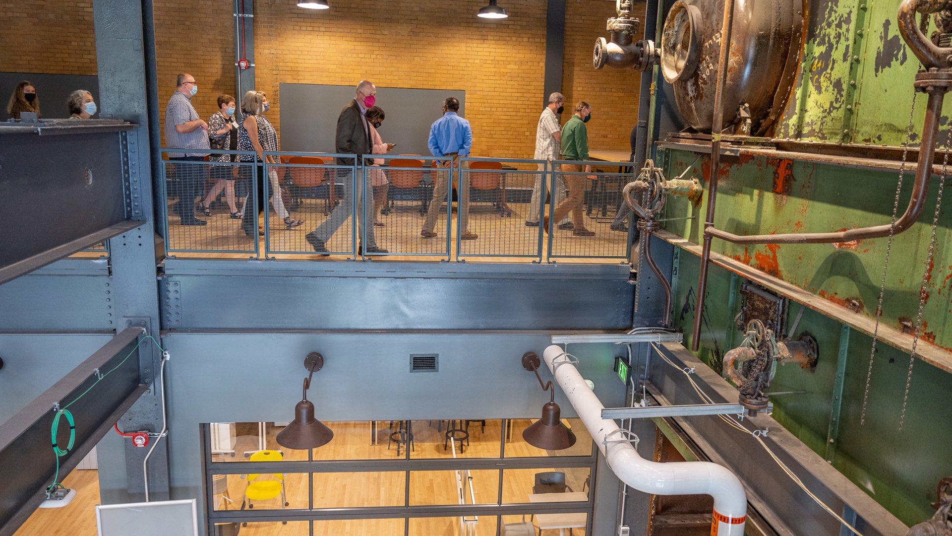 A photo shows a tour group in the new STEM Teaching Learning and Facility walking through a hall area with seating for students. From this vantage point, you can also see part of the original boiler from the Shaw Lane Power Plant.