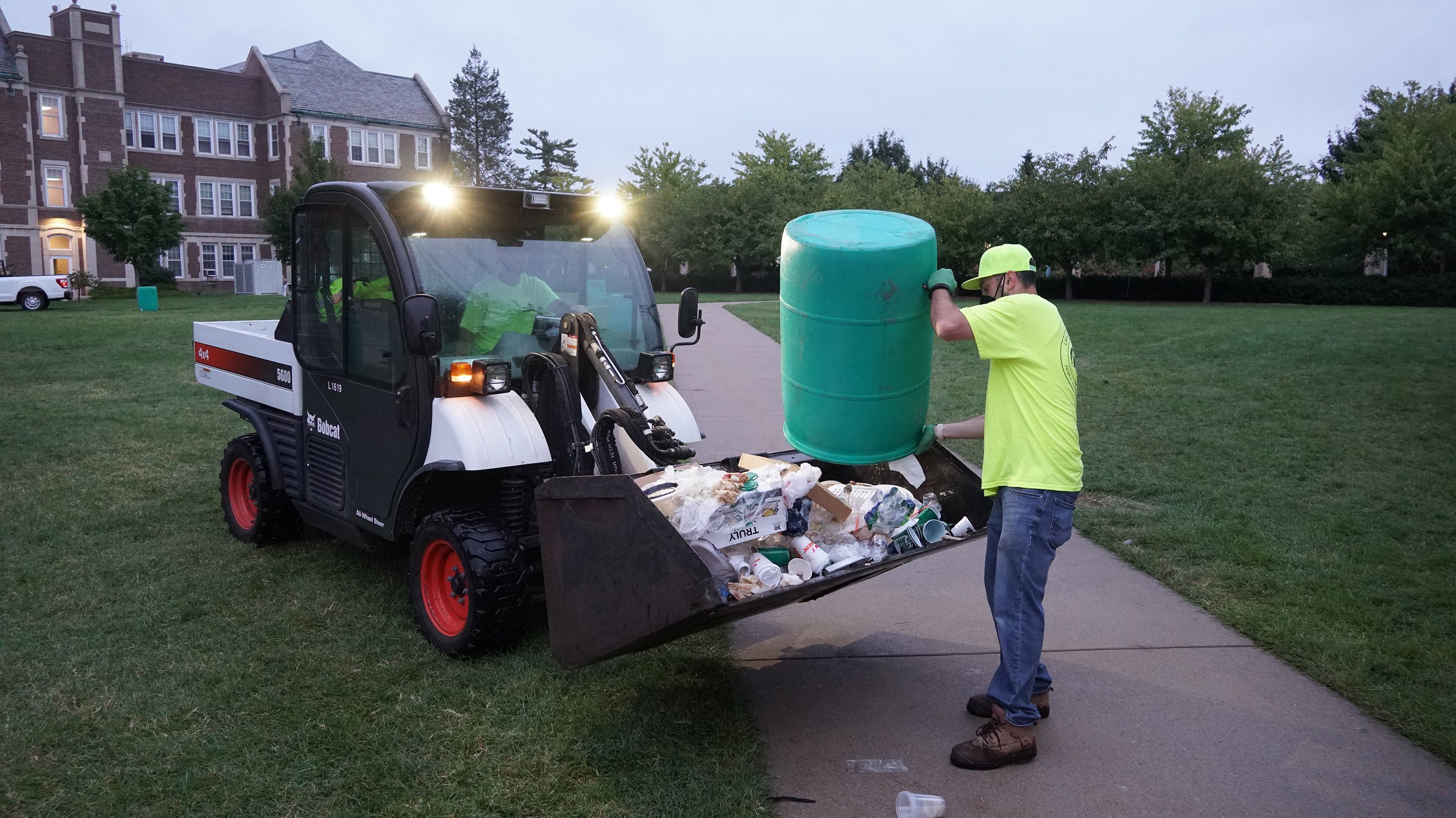 An IPF worker empties a green trash can into the scoop bucket of a Bobcat utility vehicle.