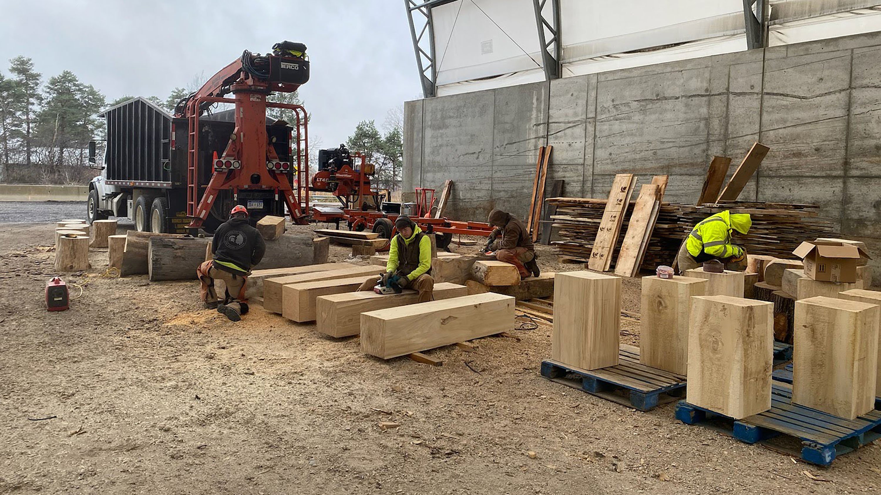 Four arborists work independently to construct outside seating and furniture out of large blocks of wood. 