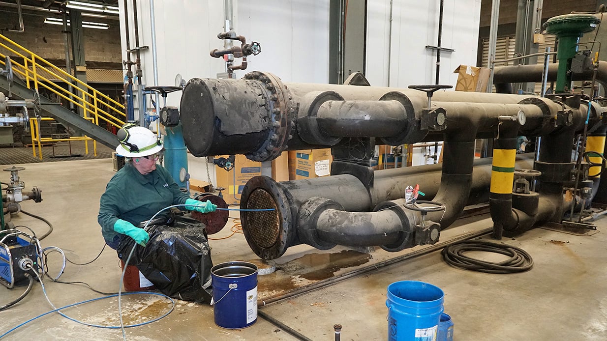 A worker in a hard hat and gloves is to the left of large iron pipes.