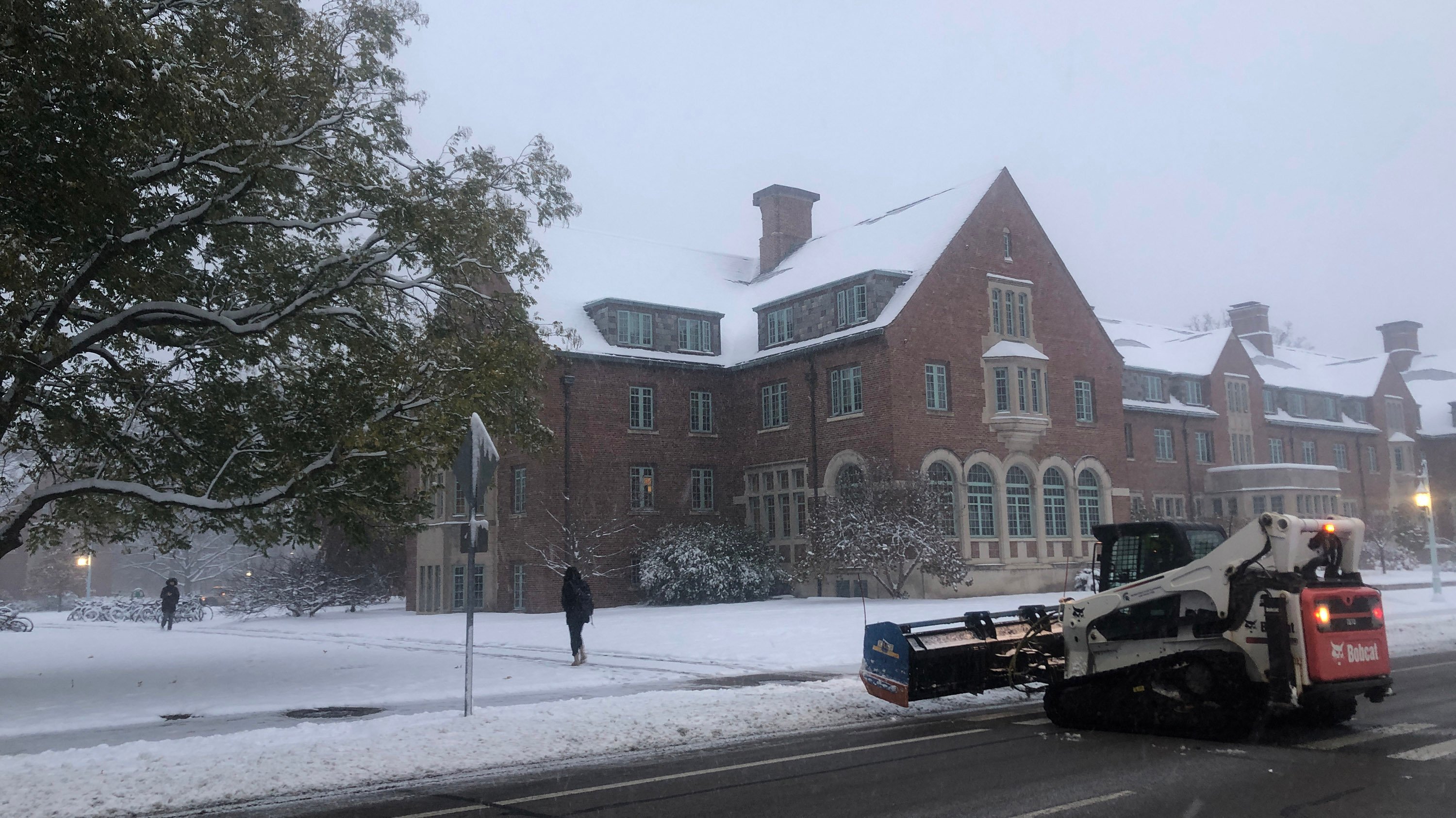 A Bobcat vehicle with snowplow attachment works to clear snow from the road with a snowy brick building in the background. 
