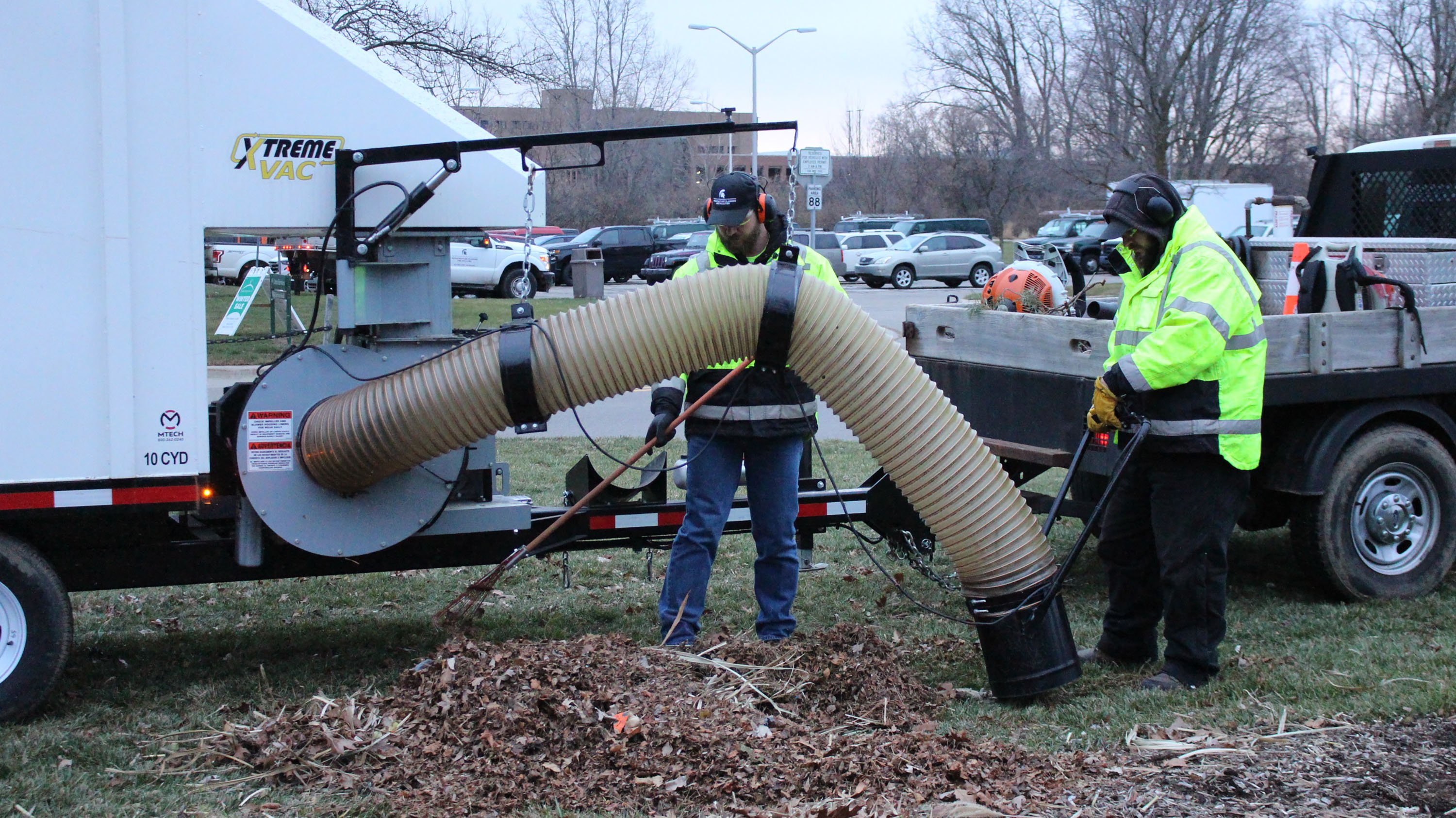 Branden Linton, a gardener, holds a rake as Mitchell Kauffman, a laborer, collects leaves and debris with an industrial vacuum hose.