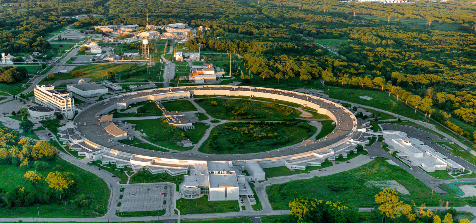 An aerial photograph looks down at the Advanced Photon Source facility, a light-colored ring-shaped building that’s more than 1,200 feet across. Other, smaller white buildings are also in the frame, dotting a lush green landscape of grass and trees.