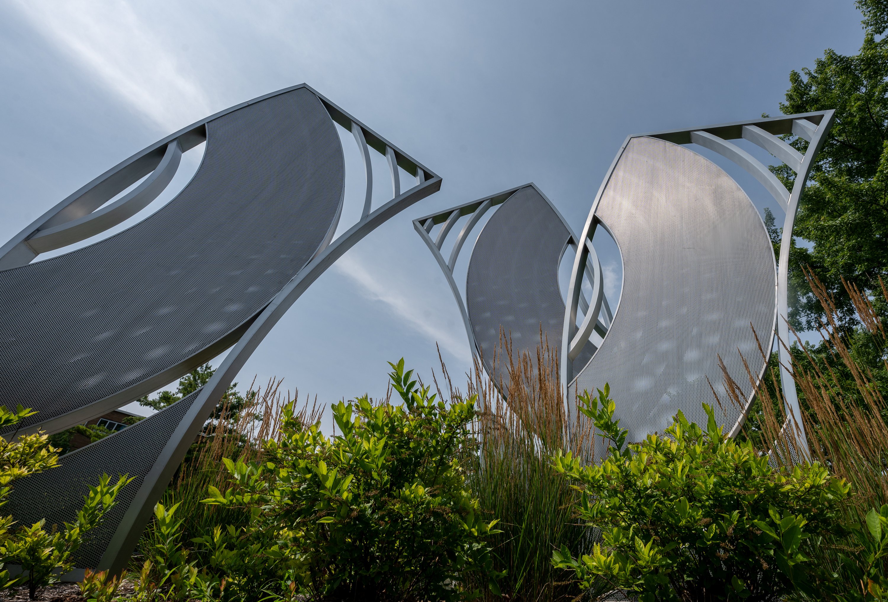 The 'ECHO' aluminum statue gleams in sunlight with green bushes in the foreground.