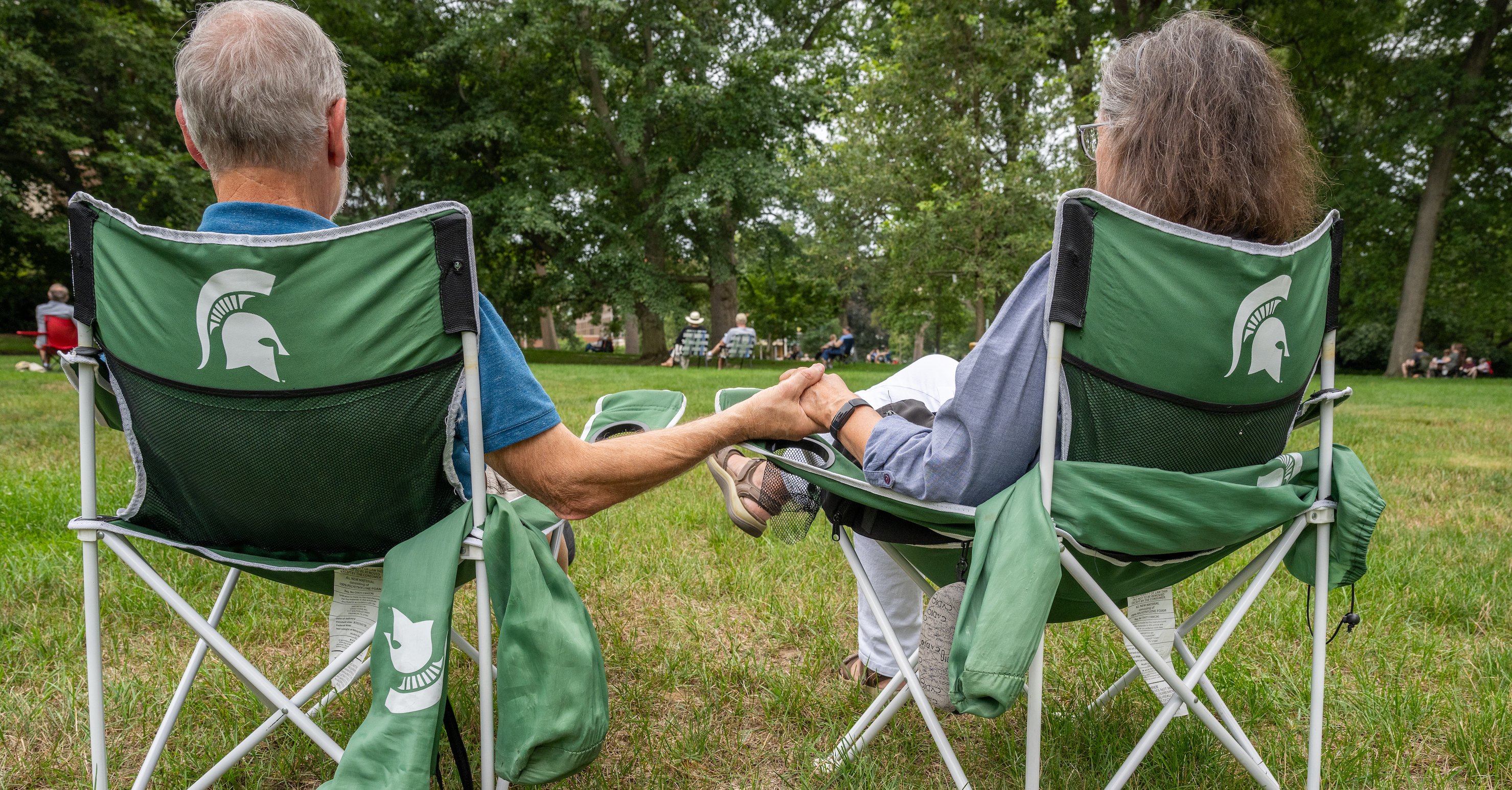 People sit in green Spartan-themed outdoor chairs, face away from the camera while holding hands. 