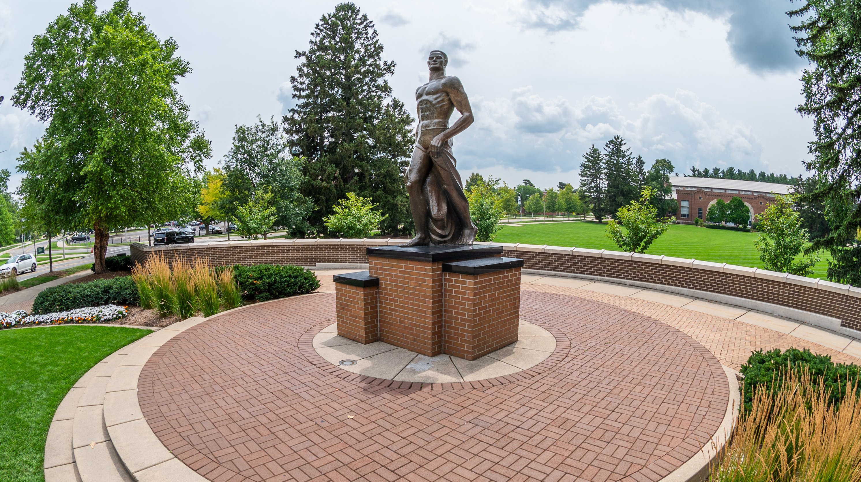 The Spartan statue stands in the middle of a paved walkway, decorated by greenery and trees.