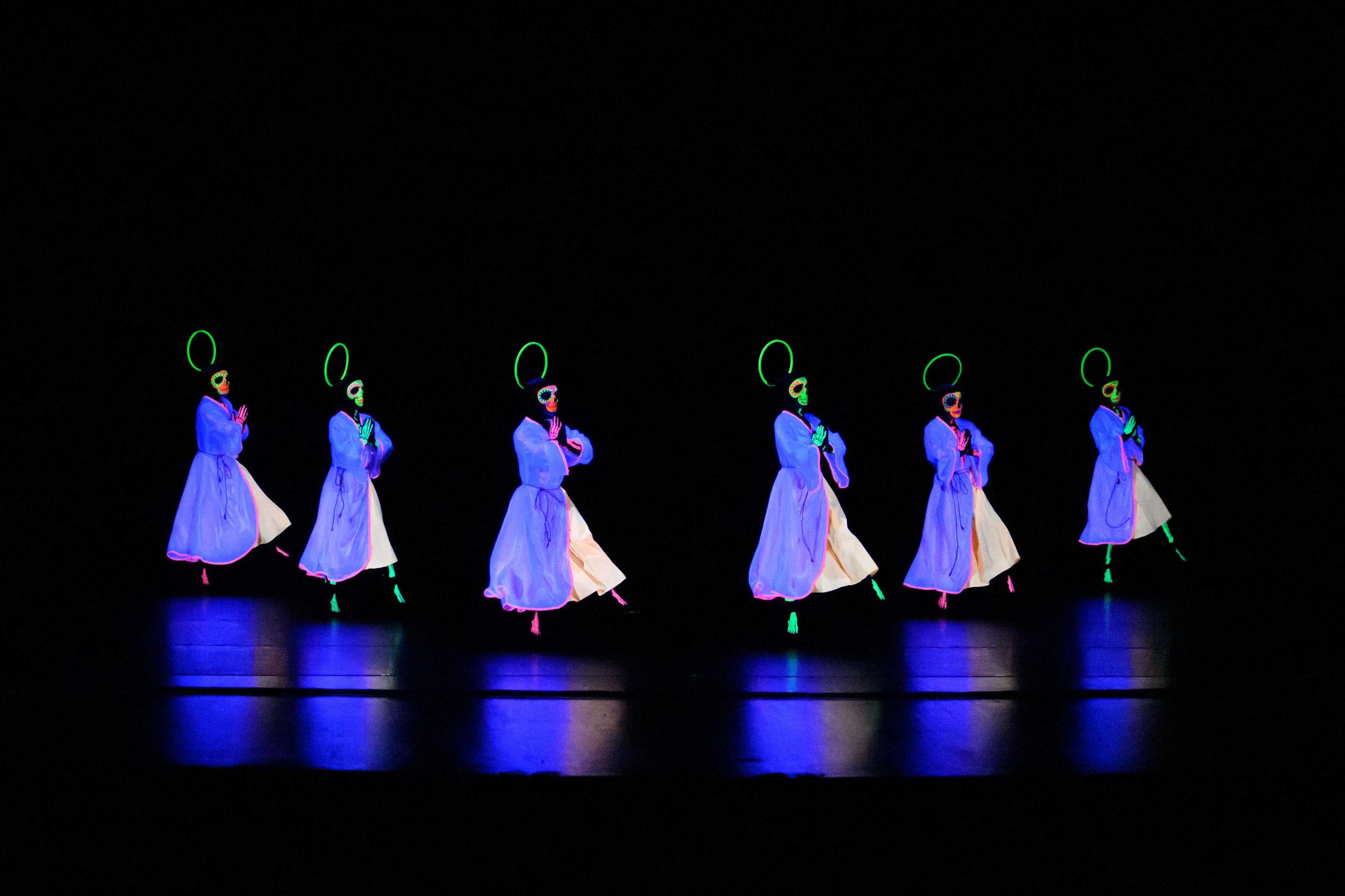 Performers in black light wearing white gowns with skeleton faces and halos