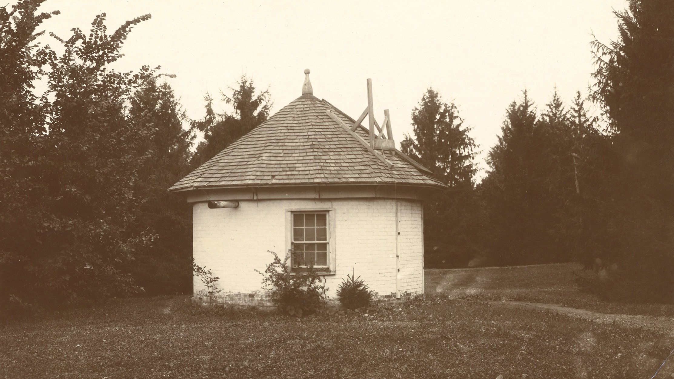 A sepia-toned historical photo of a small hut in a grass field surrounded by pine trees.