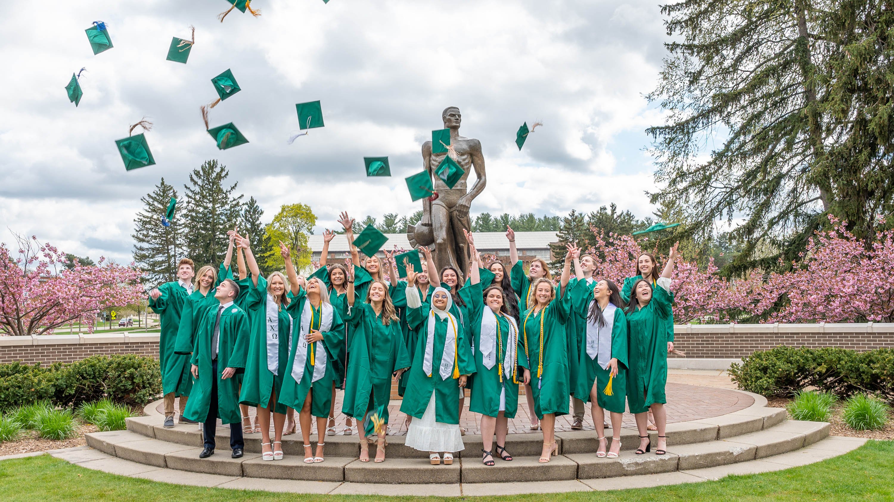 graduates tossing caps in front of Spartan statue