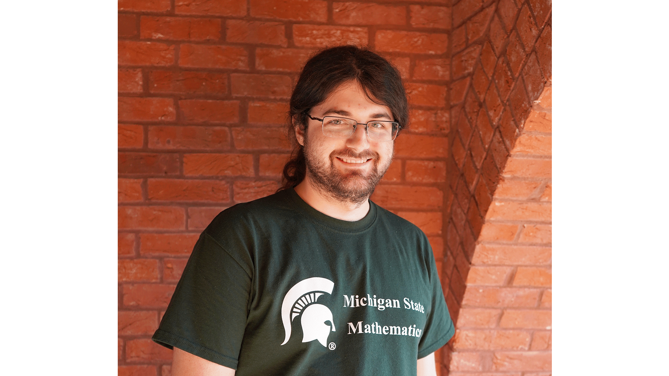 Samuel Sottile is in a green Michigan State University shirt in front of a red brick wall.