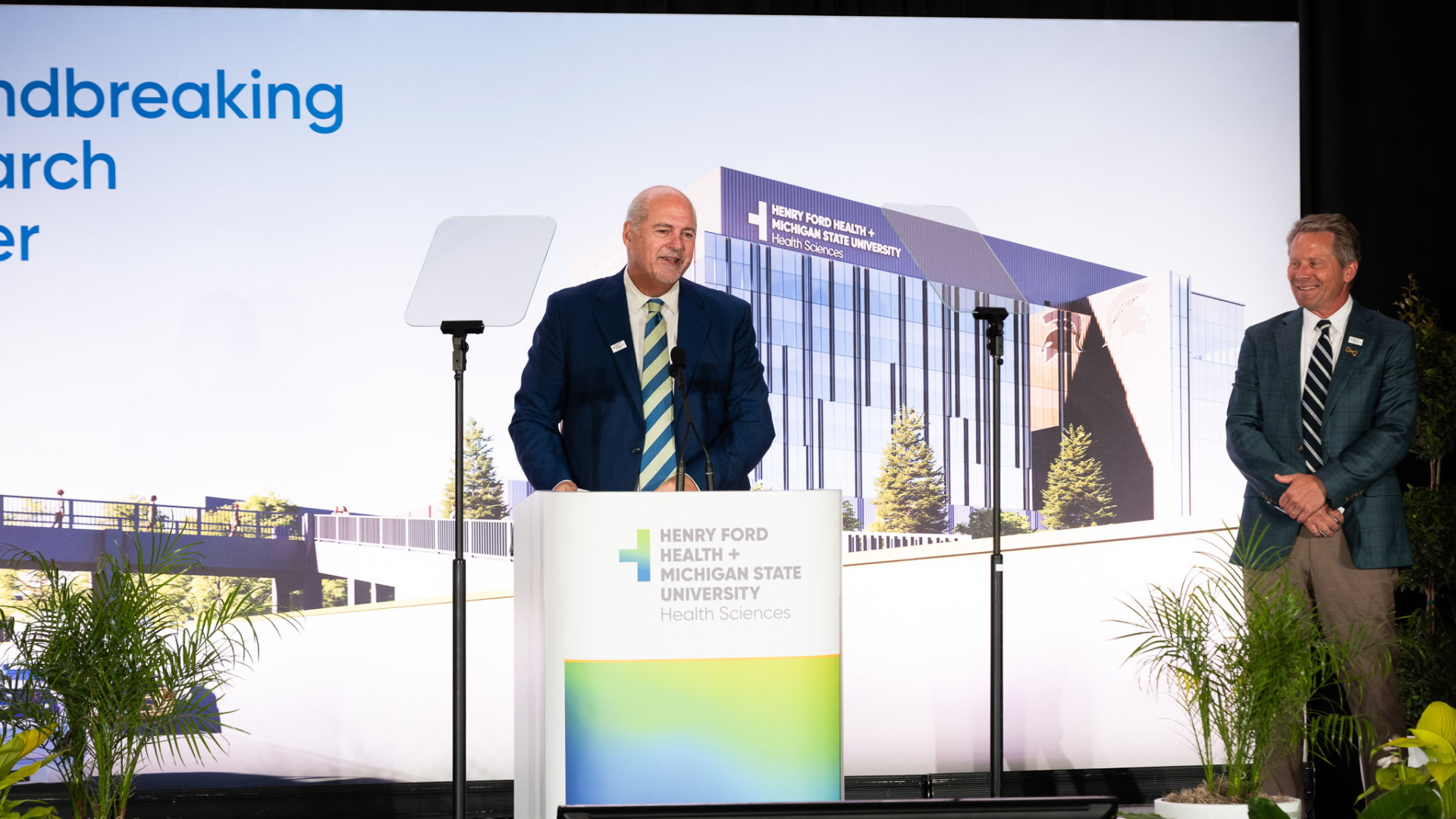 A speaker addresses the audience at the groundbreaking ceremony for the Henry Ford Health + Michigan State University Health Sciences Research Center, with a rendering of the new building in the background.