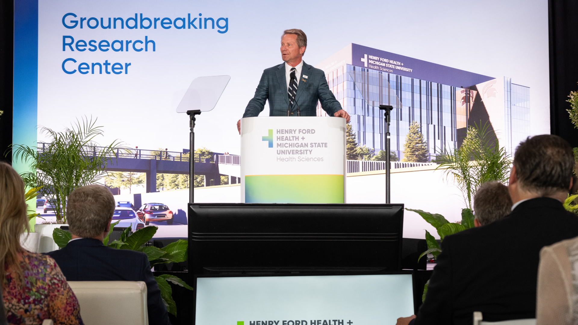  stands at the podium during the groundbreaking ceremony, with the audience seated and the new research center's design projected behind.