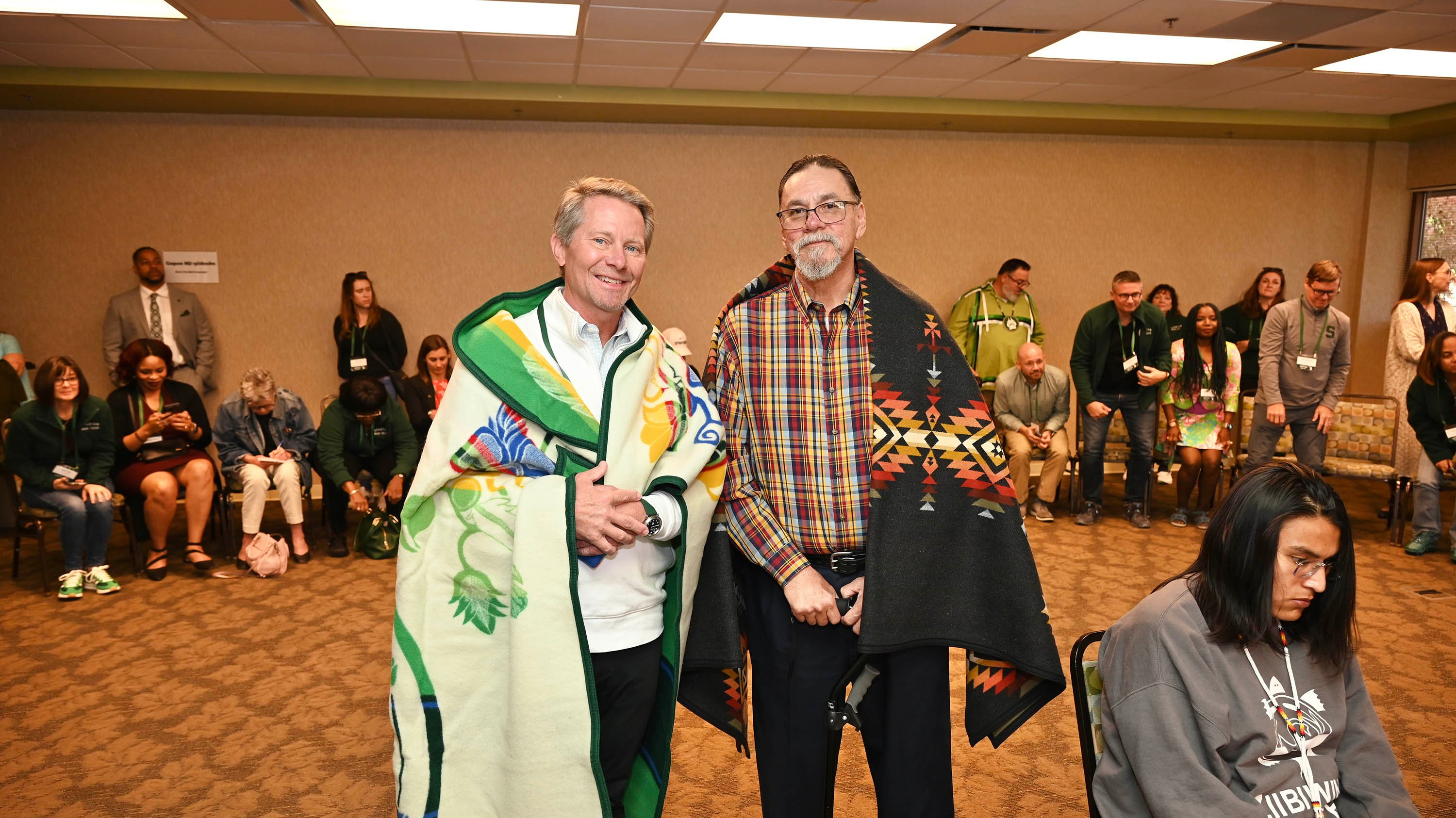 Two men, with blankets around their shoulders, pose for a photo together.