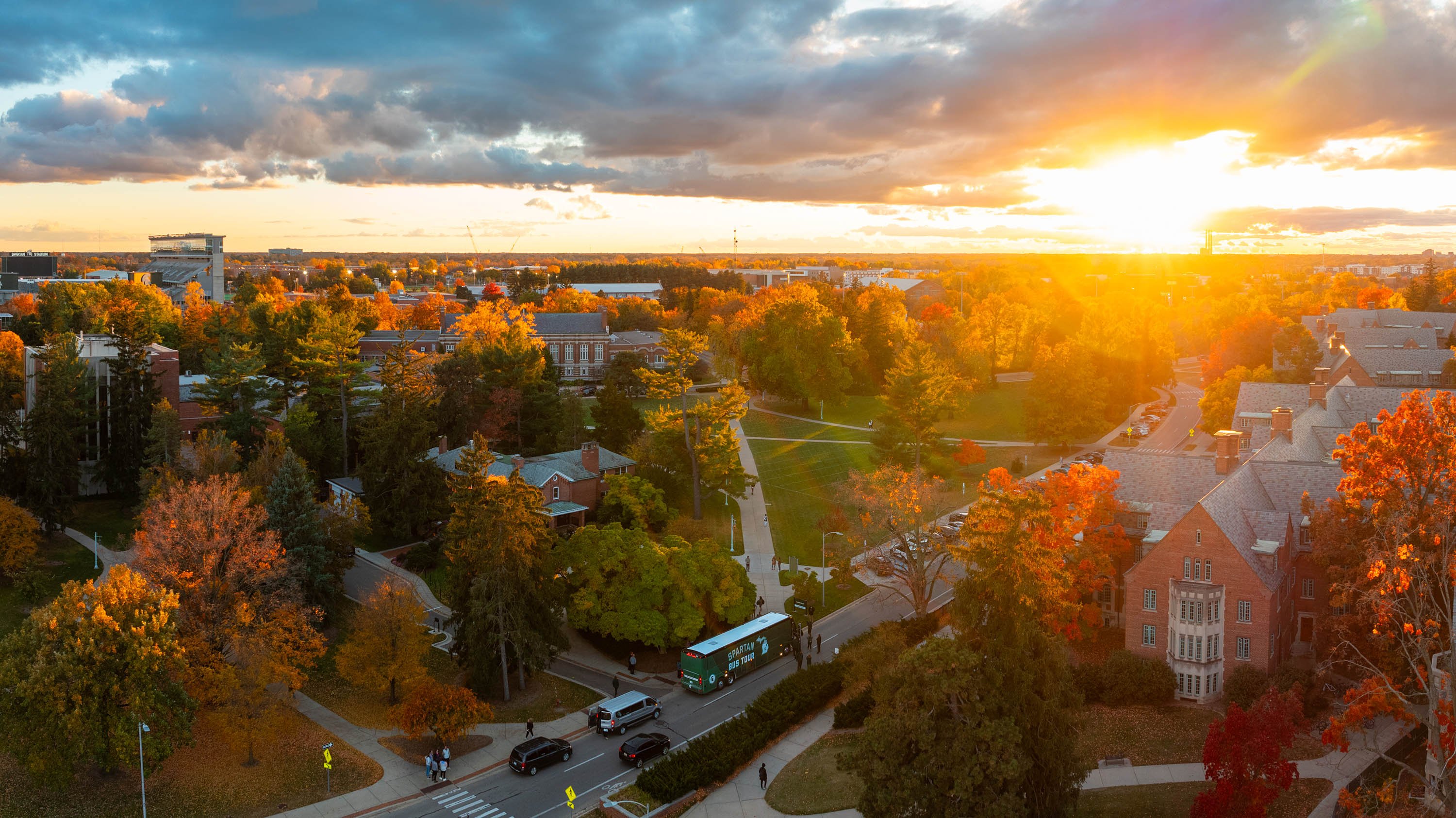 An aerial view of MSU?s campus in the fall. A green bus that says ?Spartan bus tour? in white with a white shape of the state of Michigan next to it is parked on the road.