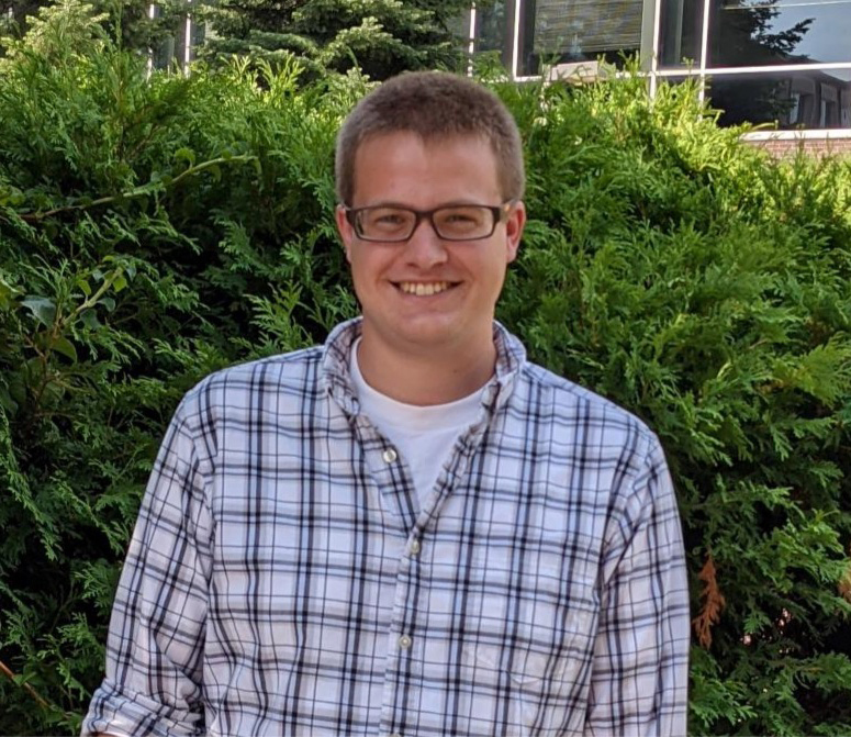 Samuel Barnett, who’s wearing in a plaid shirt, smiles at the camera. He is standing in front of an evergreen shrub.