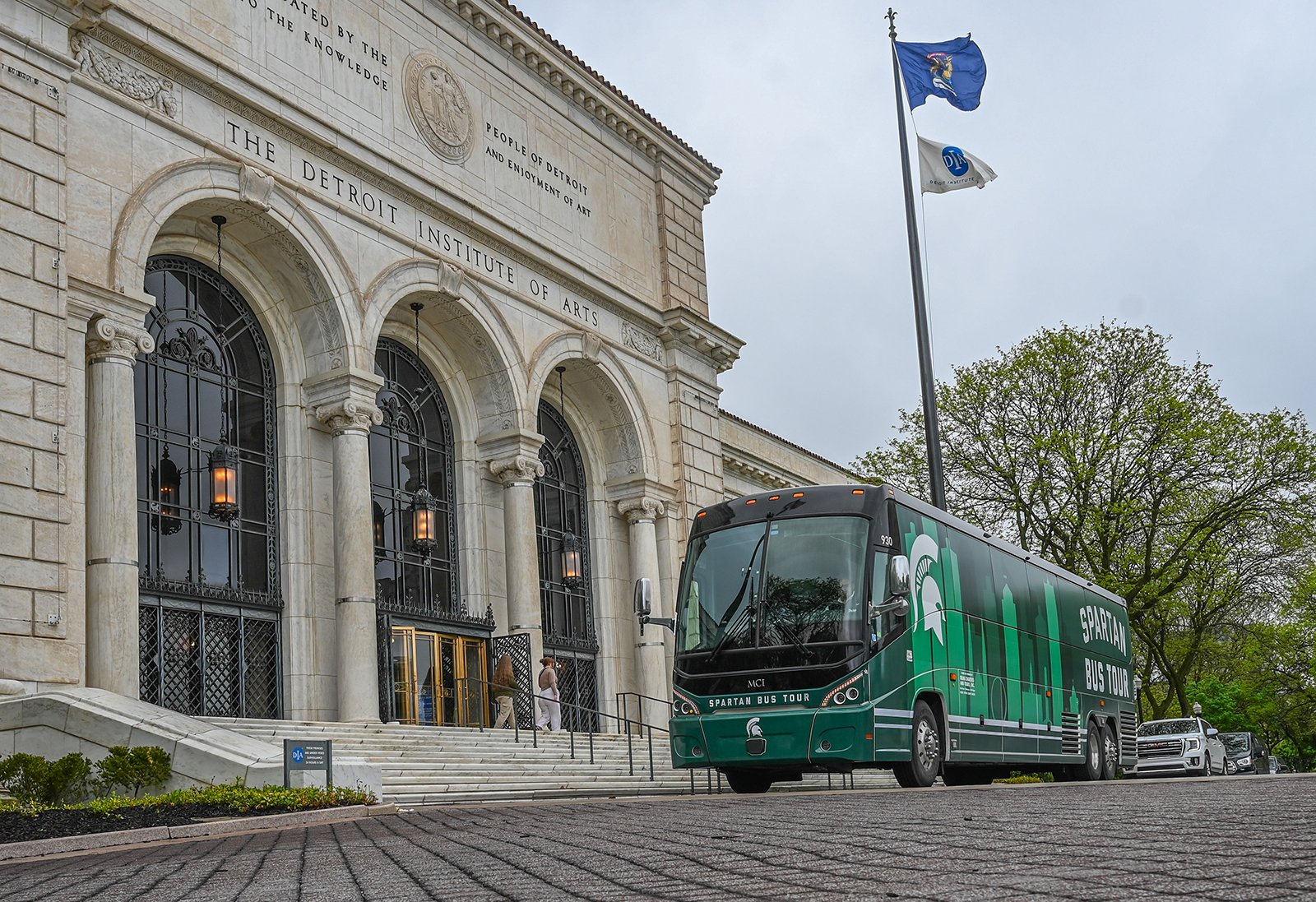 A bus with a white Spartan head and green outline of the Detroit skyline outside of a white stone building. The lettering above the entrance above the building says ?Detroit Institute of Arts?