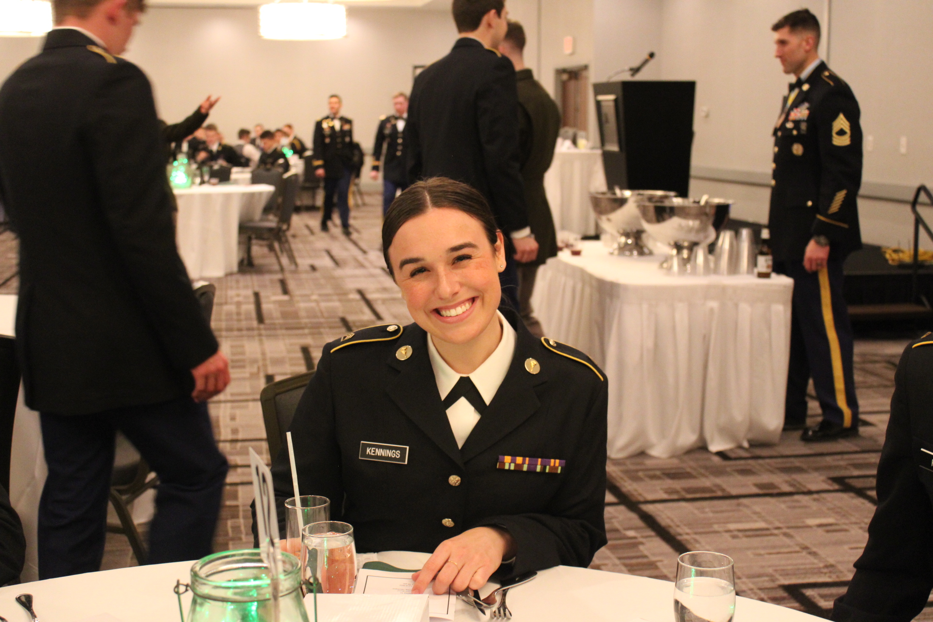 A cadet in dress uniform sits a dining table in a large dining room facing the camera and smiling.