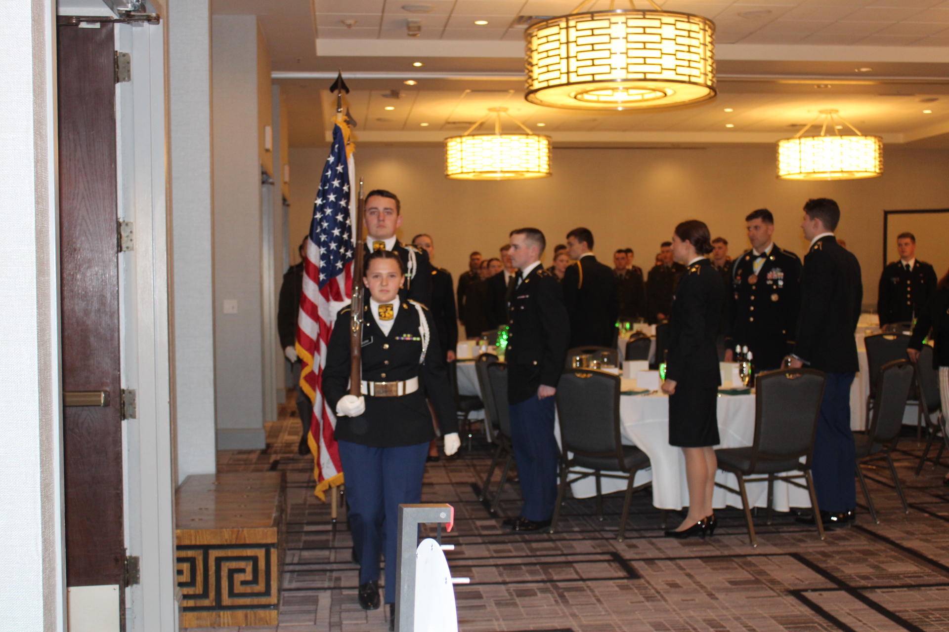 Color guard exits a banquet room where diners are standing and facing them as they exit. 
