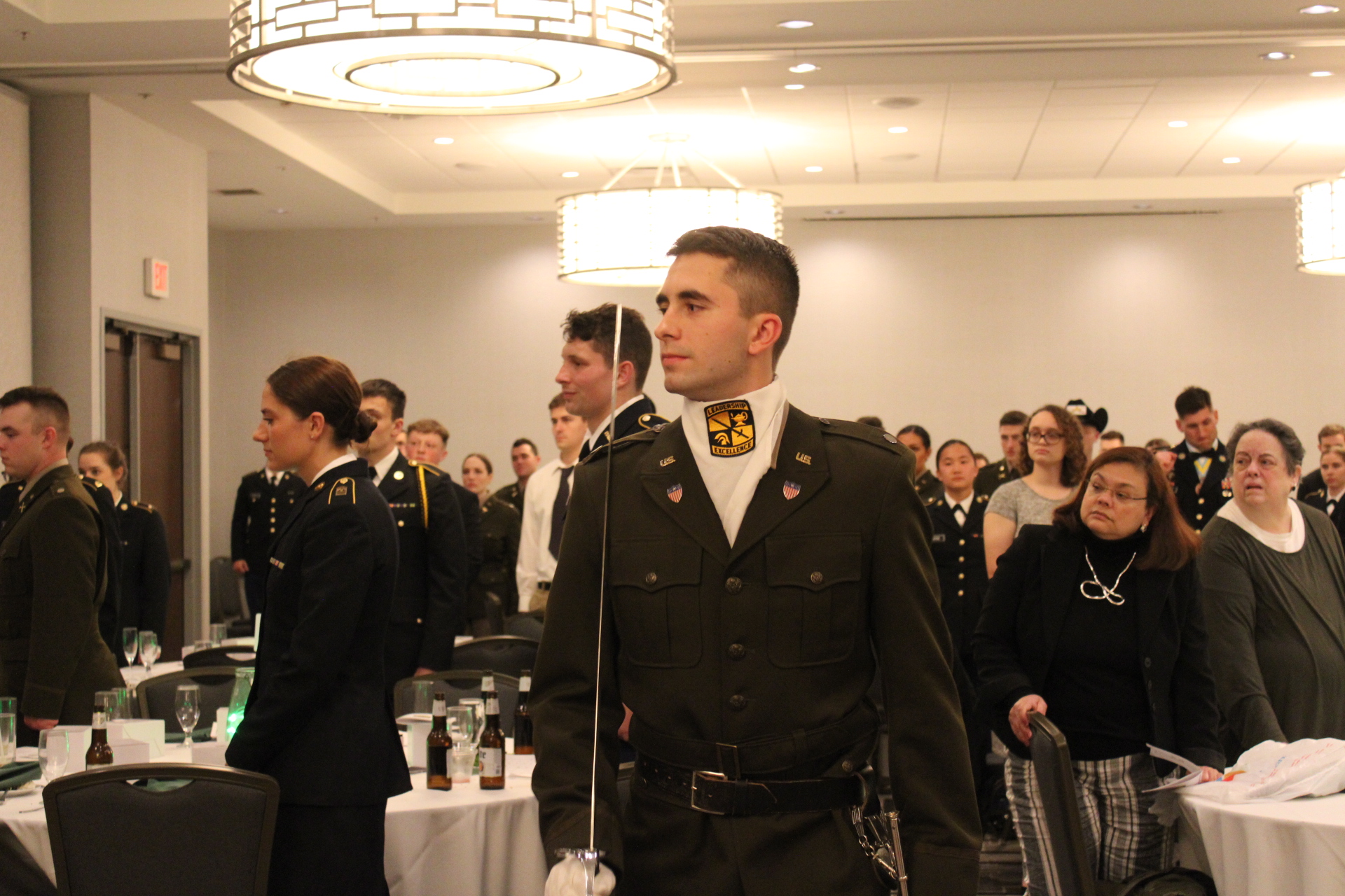 Color guard faces the front of the crowd in a banquet dining room where all the diners are standing. 