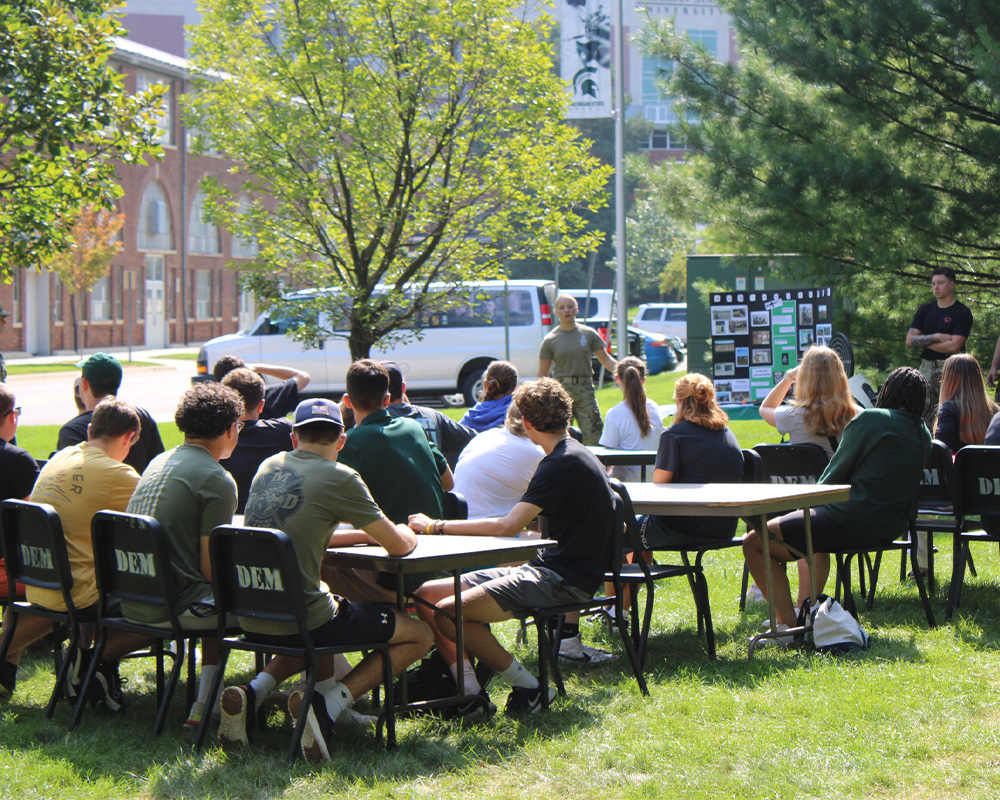 Students sit at tables outside looking at a person standing in front of a presentation board. 