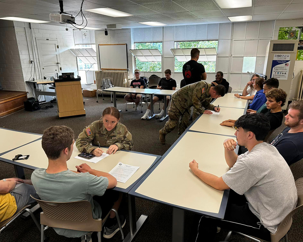 Students sit across from cadet at long tables set up in a semi circle with papers and other materials around them.