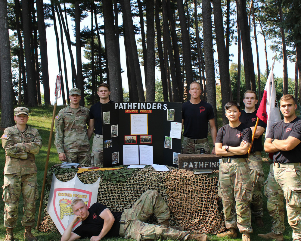 A group of cadets face the camera while gather around a pathfinders table set up. 