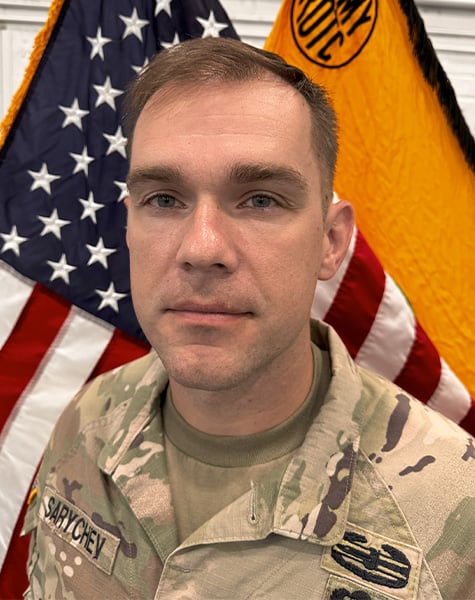 Portrait. Subject is featured from mid-chest up, standing in front of an American flag and yellow flag. They are wearing Army fatigues with hair cut short. 