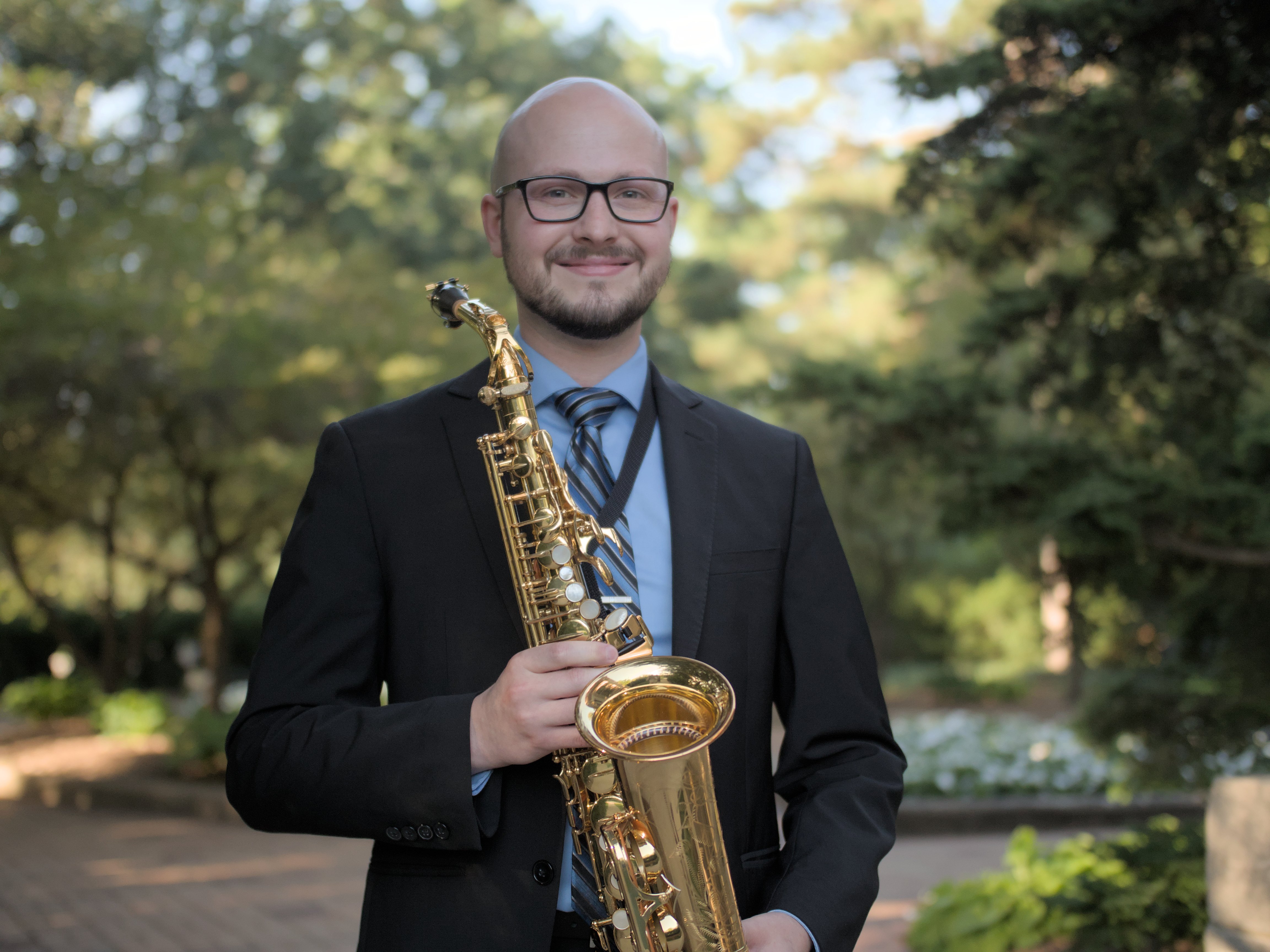 man with glasses holding a saxophone standing in a garden