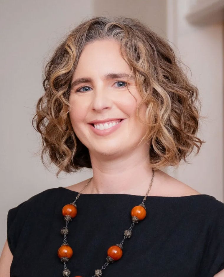 Lauren Klein, wearing a black blouse and a metallic necklace with orange beads, standing in front of a beige background.