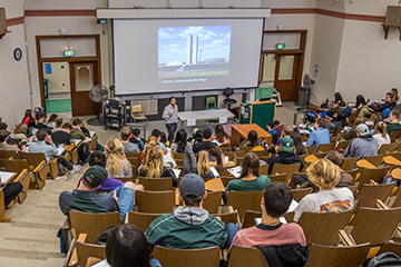 Students sit in a full lecture hall with a professor lecturing in front of a slideshow presentation.