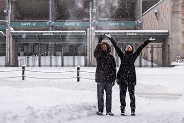 Two people stand outside Spartan Stadium in the snow as snow falls around them.