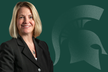 Headshot of Teresia Hagelberger, Director of Faculty and Academic Staff Affairs, smiling and wearing a black jacket in front of a green background featuring a Spartan helmet logo.