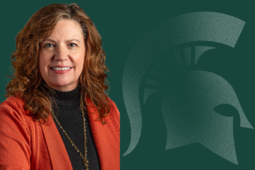 Jennie Schaeffer, Director for Faculty and Academic Staff Affairs, smiling, wearing a red jacket over a black blouse standing in front of a green background with a Spartan logo.