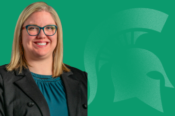 Kara Yermak, Assistant Provost for Faculty and Academic Staff Affairs, smiling, wearing a black jacket and blue blouse, standing in front of a green background featuring a Spartan helmet logo. 