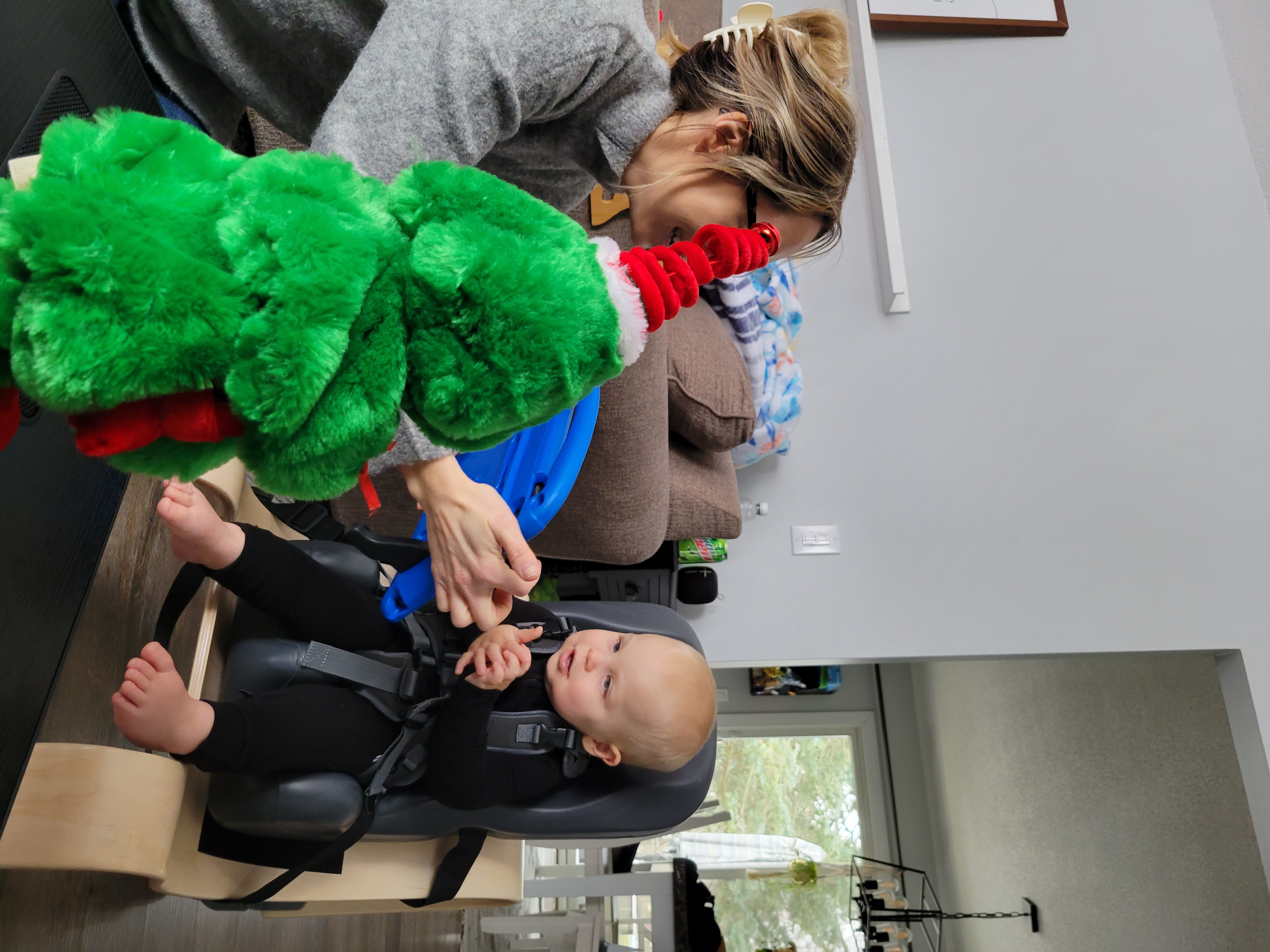 Baby in car seat with woman buckling them in. Green dinosaur stuffy in forefront 