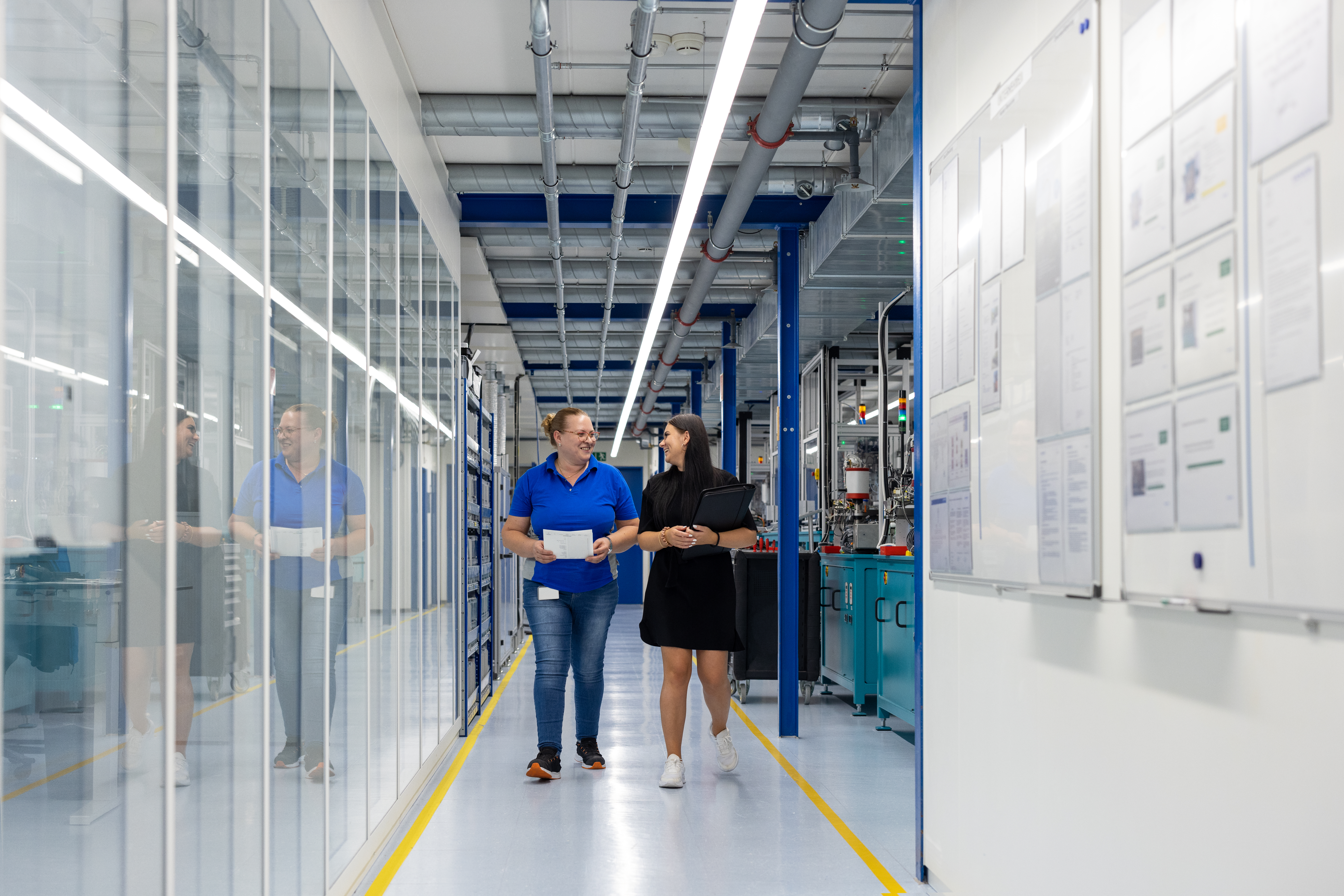 Two people walk down a hallway of glass windows in a manufacturing building. 