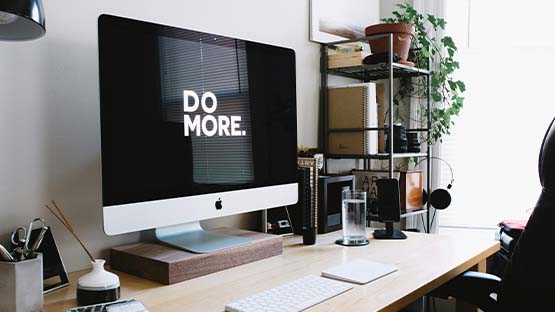 A workspace containn a light wood desk with shelves, plants, various work accessories and an Apple computer monitor displaying "DO MORE."