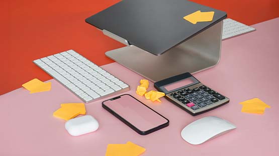 A photograph of a closed MacBook on a metal laptop stand, surrounded by a keyboard, phone case, mouse and calculator. Orange arrow-shaped PostIt notes point between the items to illustrate workflow.