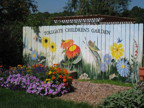 A fence painted with flowers and bugs with flowers blooming in front of it