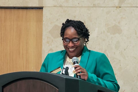 Dr. Ebony Green stands at a podium with a microphone in her hand, while she smiles and looks down at what is in front of her. She is wearing a green blazer, with dark, short curly hair. 