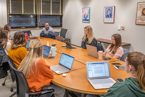 A group of students with laptops open sit around a wooden oval table with their instructor speaking at the head of it