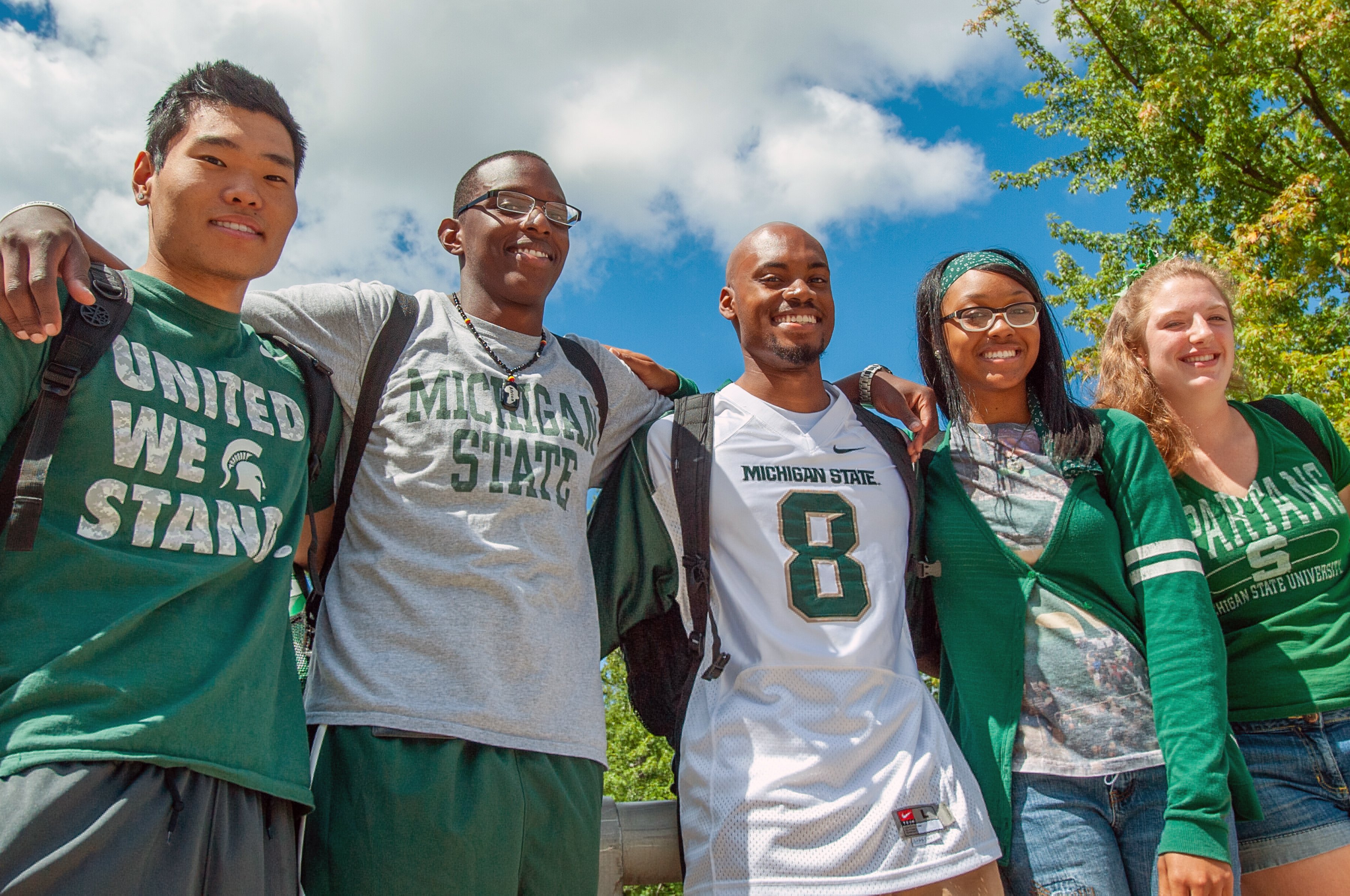 A group of Spartans wearing MSU brand clothing stand smiling with arms around each other on a bright summer day.