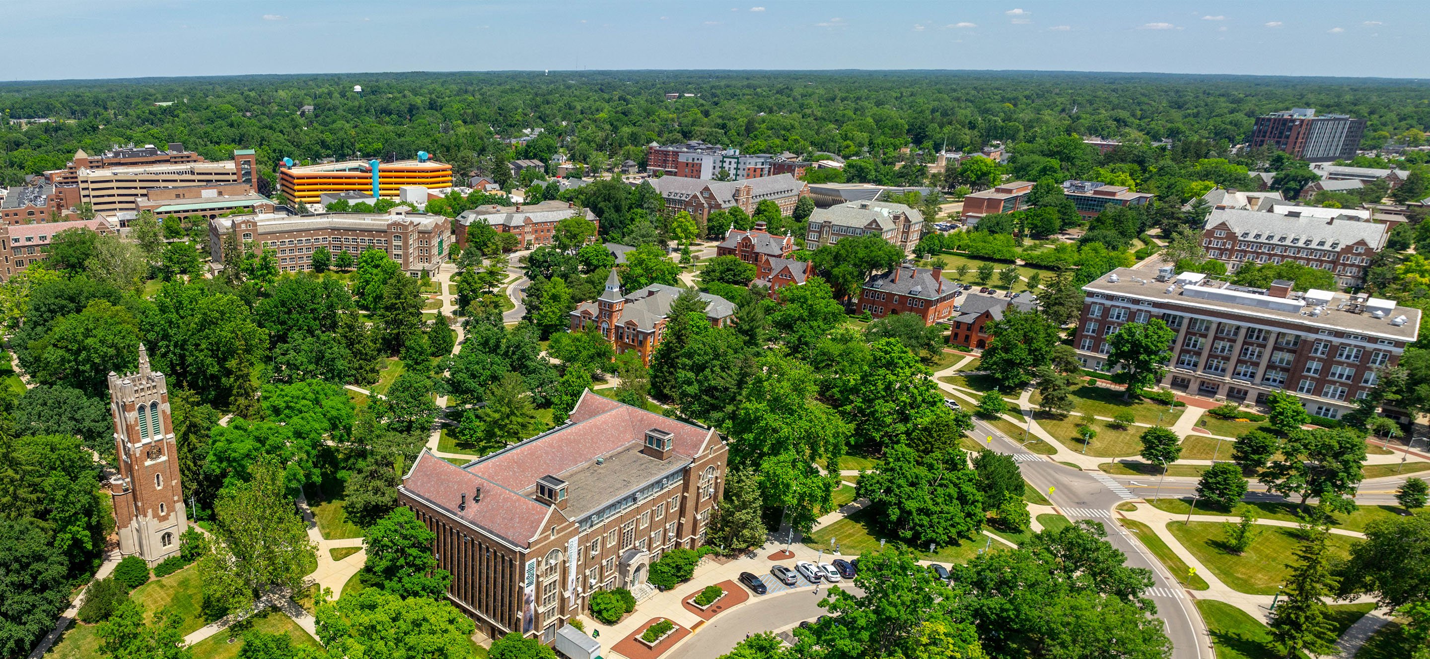 Aerial view of MSU's campus with red-brick academic buildings, walking paths, and roads arranged around open green spaces, stretching to a forested horizon.