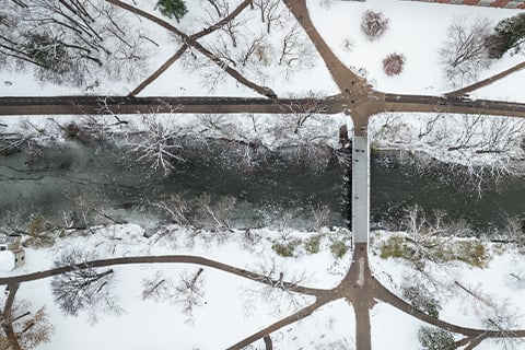 Aerial view of a snow-covered campus landscape with intersecting walking paths, leafless trees, and a narrow river running through the center crossed by a pedestrian bridge.
